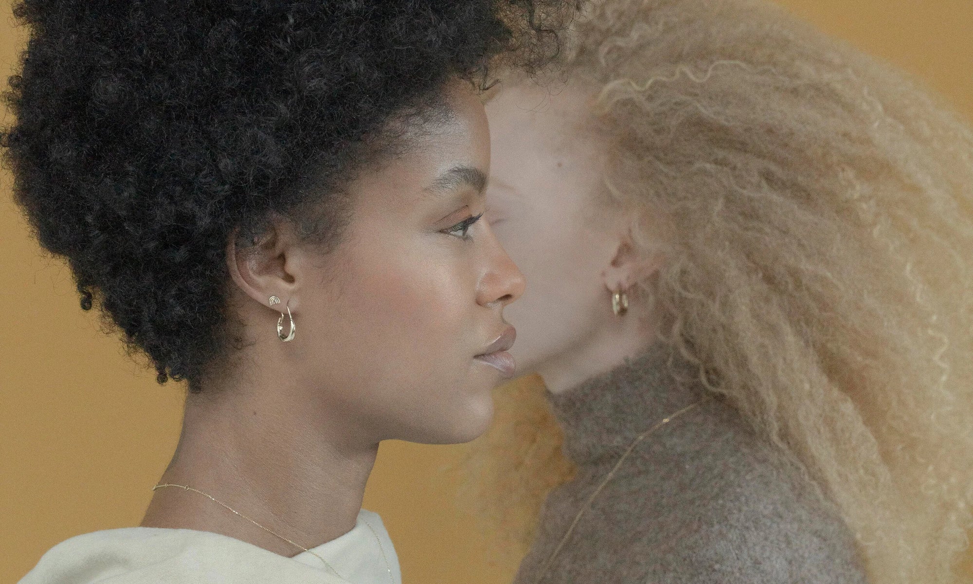 Models Alicia Myers and Nyasia Wyani stand opposite in the Talisman Fine Jewelry Alchemy Collection. From left, Alicia wears the Mini Rainbow Stud and Aphrodite Hugs; Nyasia wears the Rollo Hoops and U Earrings. All sustainably handmade in recycled 14k gold, using traditional lost-wax casting techniques.