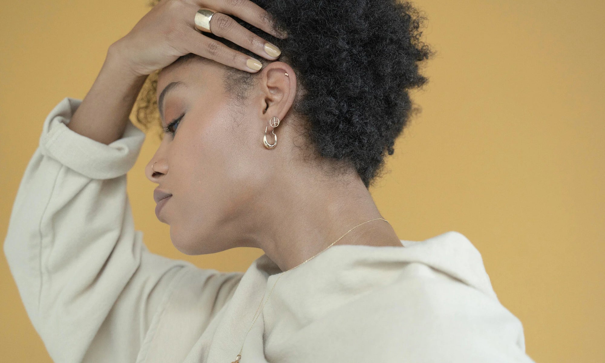 Model Alicia Myers wears the Aphrodite Hugs with the Peace Sign Studs and Mini Orb Studs, along with the Demeter Cigar Band. All sustainably handmade in recycled 14k gold.