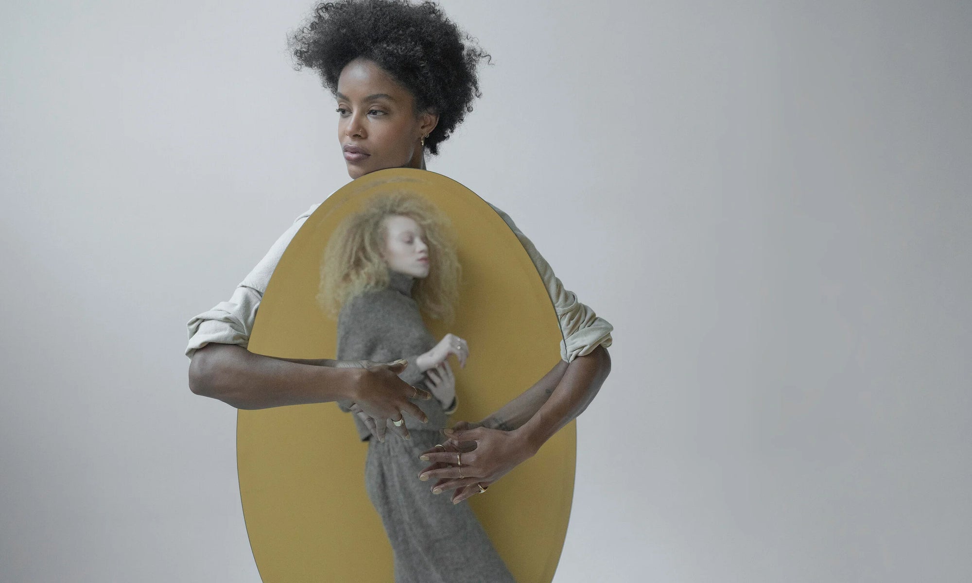 Model Alicia Myers holds Nyasia Wyani in her reflection, both wearing the Talisman Fine Jewelry Alchemy Collection. Clockwise from top right: Aphrodite Hugs, Peace Sign Studs, Ra Pinky Signet, Circle Shape Ring, Rollo Band, Triangle Shape Ring, Demeter Cigar Band. All sustainably handmade in recycled 14k gold, using traditional lost-wax casting techniques.