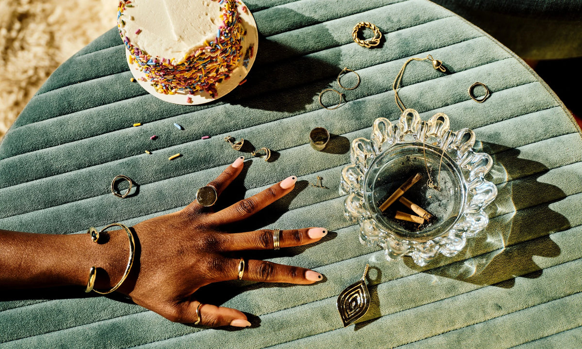 Model Samantha wears the Inverse Cuff, the Portal Bracelet, Ra Pinky Signet, Infinite Ring Stack, Portal Ring, and the Wave Theory Ring. On the ottoman, clockwise from top left: the Shapeshifter Ring, Held Earrings, Demeter Cigar Band, Infinite Hoops, Helix Ring, Shroom Necklace, Sirius B Ring, Wave Earring, and the Mary Pendant. All sustainably handmade in solid, recycled 14k gold.